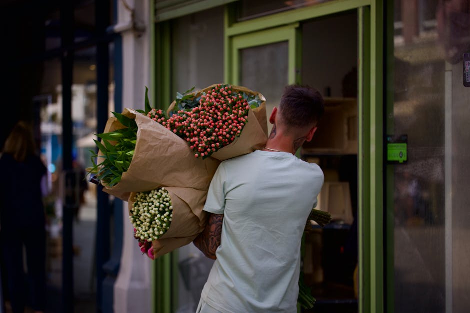 A person with short dark hair, wearing a light-colored T-shirt, is seen from behind holding a large bouquet of fresh flowers wrapped in brown paper and tied with white ribbon. The bouquet includes vib