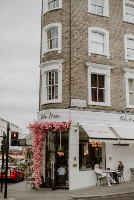 A floral arrangement featuring a cascading display of soft pink blossoms, likely cherry blossoms or similar flowers, adorns the corner of a white storefront on a street corner. The flowers drape elega