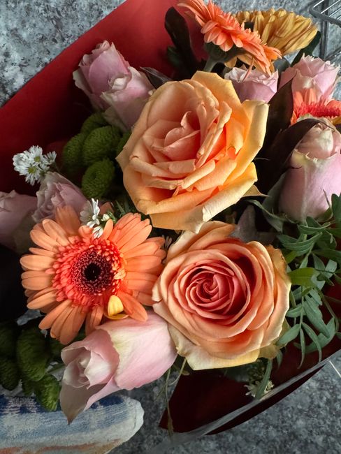 A floral arrangement featuring a mix of roses, gerbera daisies, and spray roses in shades of peach, pink, and soft orange, accented with green foliage. The roses have full, layered petals with a velve