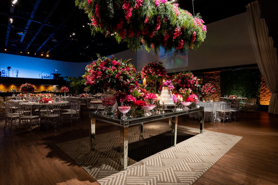 A luxurious event space with a dark wooden floor and a black ceiling, decorated with an abundance of vibrant pink floral arrangements. In the center, a mirrored rectangular table holds large bouquets 