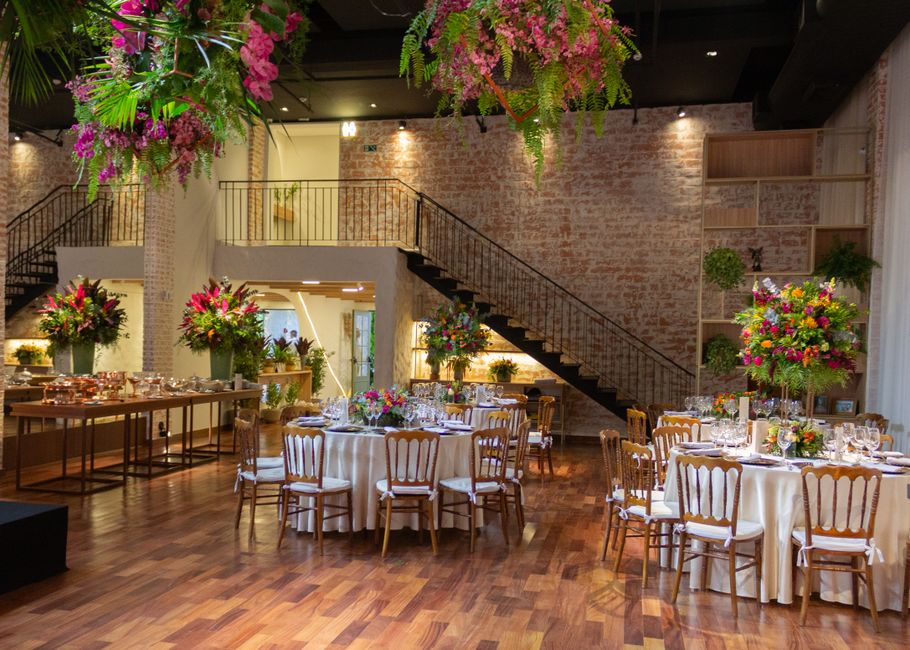 A spacious interior of an event venue decorated for a celebration, featuring multiple round tables draped in cream tablecloths and adorned with elegant floral centerpieces comprising pink, purple, red