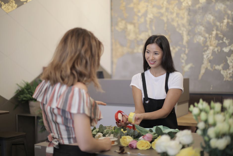 A professional florist wearing a black apron over a white t-shirt is handing a floral gift wrapped in yellow paper with a red ribbon to a customer with shoulder-length blonde hair and a striped blouse