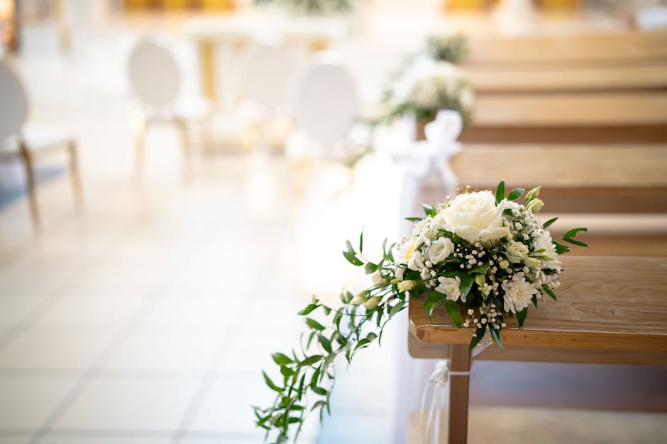 A delicately arranged bouquet of fresh white flowers, including roses and baby's breath, with lush green foliage, resting on the edge of a polished wooden pew in an indoor ceremony setting. The bouque
