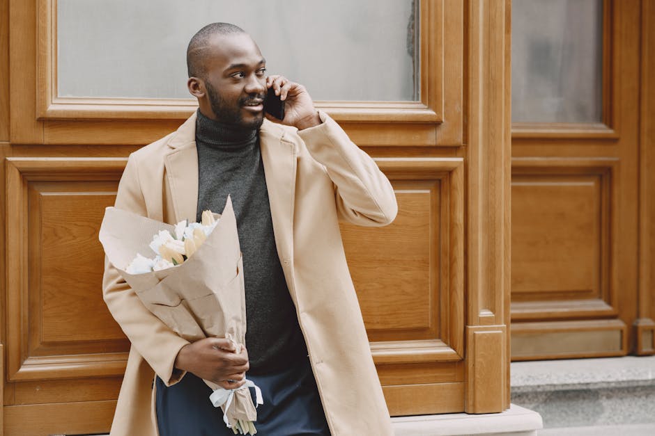A man with dark skin and a beard is standing outdoors in front of wooden panel walls, holding a bouquet of fresh white roses wrapped in brown craft paper. He is talking on a mobile phone, with a sligh