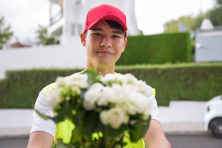 A delivery person wearing a red cap and high-visibility vest presents a bouquet of fresh white flowers, including roses and lilies, wrapped in clear cellophane with green accents. The person's face is